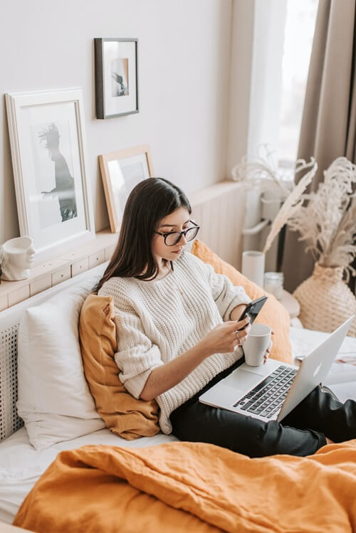 Photo d'une femme assise sur un lit, un téléphone et une tasse dans la main et un ordinateur portable sur ses genoux