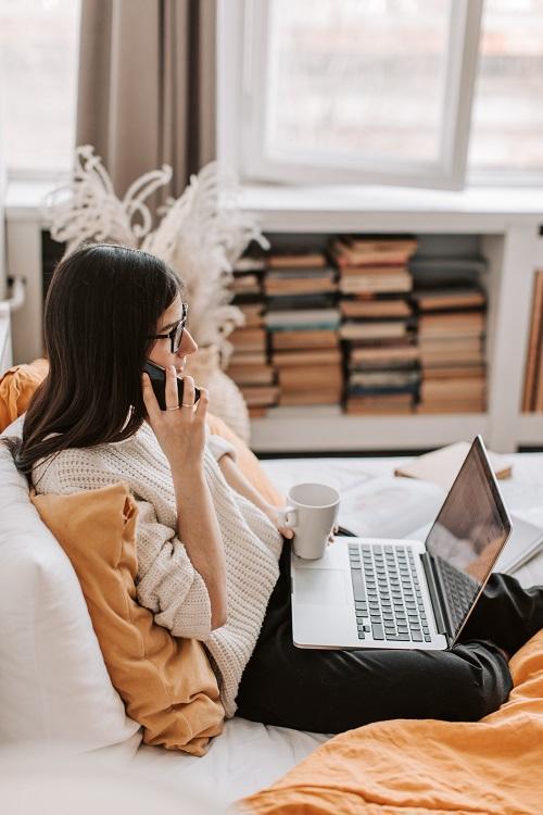Photo d'une femme assise sur un lit, un téléphone et une tasse dans la main et un ordinateur portable sur ses genoux.