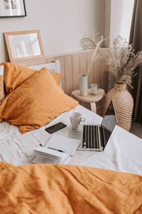 Ordinateur portable posé sur un lit avec un téléphone, un carnet et une tasse.